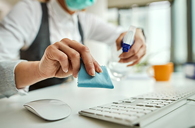 A person wearing a mask and gloves is cleaning a white desk in an office environment. They are using a blue cloth and spray cleaner, highlighting attention to hygiene and cleanliness in professional settings.